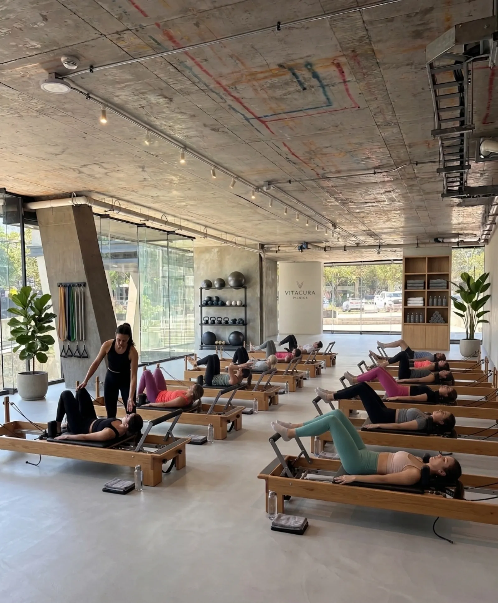 Clase de pilates con máquinas reformer en el local, bajo el cielo de hormigón a la vista y ventanal a la calle
